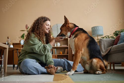 Side view portrait of happy young woman giving treats to pet dog while sitting on floor in cozy home together photo