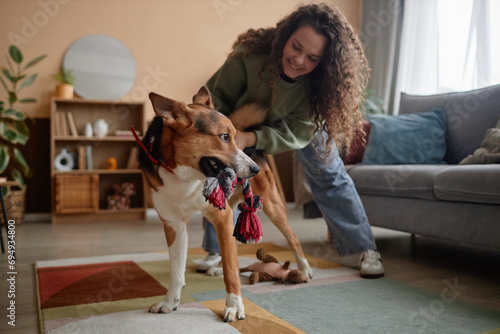 Full length portrait of carefree young woman playing with big dog in cozy living room at home, copy space