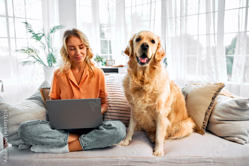 Freelance and pet. Beautiful caucasian woman with blond hair using wireless laptop on couch with golden retriever sitting near. Domestic animal feeling relaxed beside working female owner at home.