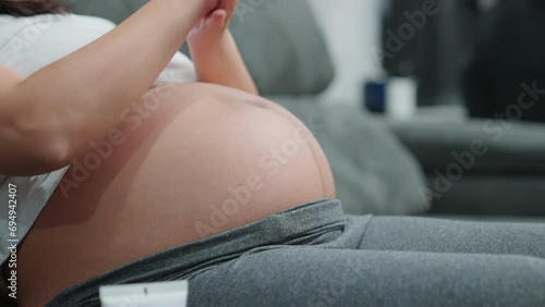 Pregnant woman applying skincare on abdomen and massages at home. Closeup belly mother rubbing gently moisturizer cream on tummy skin indoors. Taking care of mother's skin health before giving birth.