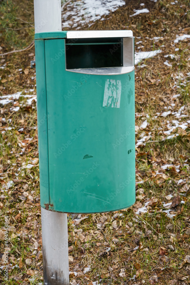 Green trash can on metallic pole with grass and snow background