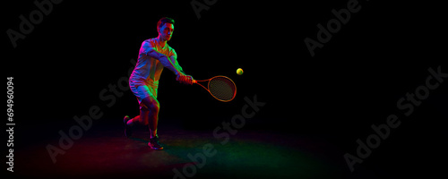 Competitive man, tennis athlete in motion during game, playing, practicing against dark background in neon light. Concept of professional sport, competition, game, math, hobby, action