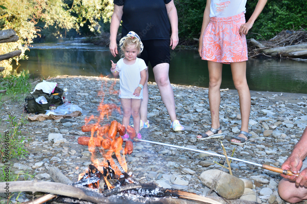 The family is camping by the river and roasting sausage over a natural fire