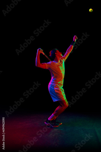 Full-length of man in his 30s, tennis player during game, hitting ball with racket against dark background in neon light. Concept of professional sport, competition, game, math, hobby, action