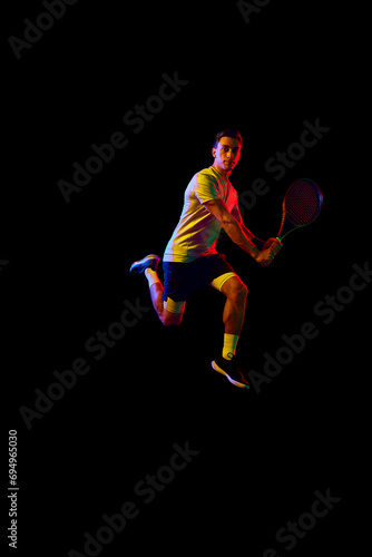 Dynamic image of man, tennis player in motion with racket against dark background in neon light. Concept of professional sport, competition, game, math, hobby, action