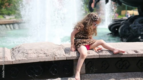 Little barefoot girl sits near fountain and swings legs