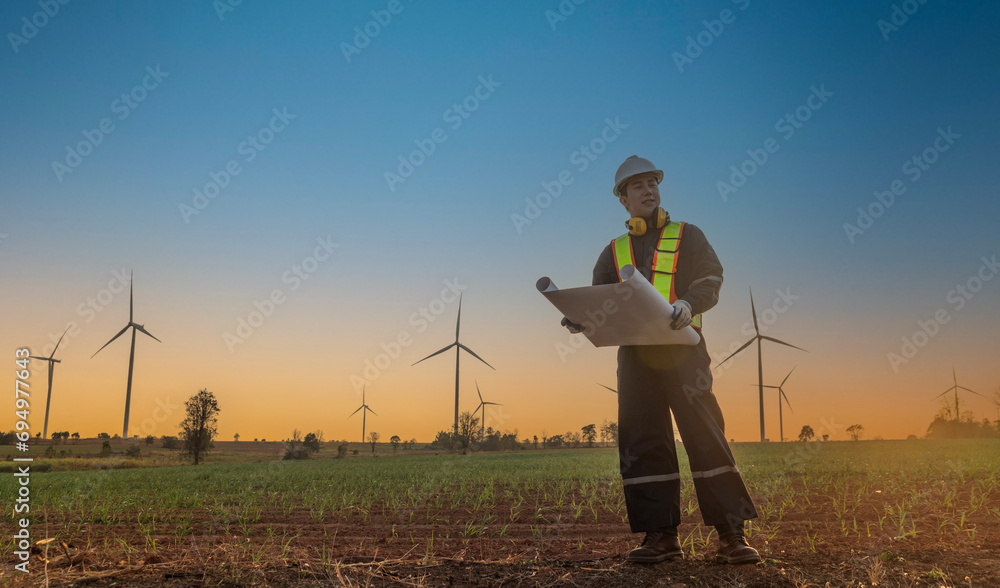 Asian male engineer standing in a turbine field holding blueprints inspecting a wind turbine generator station to keep electricity running until the sunset. Asian male engineer standing in a turbine field holding blueprints inspecting a wind turbine generator station to keep electricity running until the sunset.