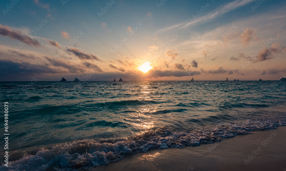 Amazing colors of tropical sunset. Sail boats silhouettes floating on ocean horizon. Ocean waves wash the sand. Boracay island, Philippines.