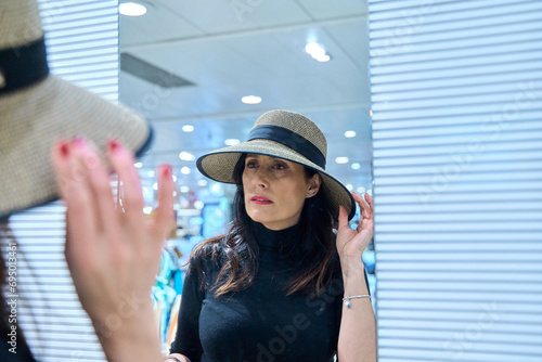 Mature woman standing in front of mirror , putting on a hat. brunette woman trying on hats in a shop to buy fashion accessories.