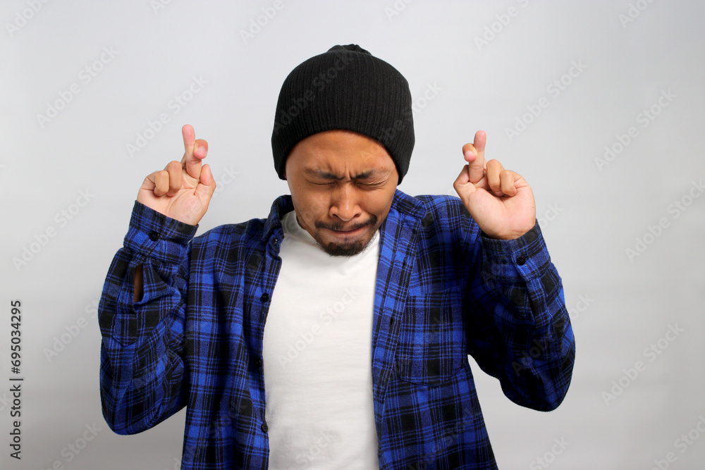 Hopeful young Asian man, dressed in a beanie hat and casual shirt, makes a wish while crossing his fingers, standing against a white background Hopeful young Asian man, dressed in a beanie hat and casual shirt, makes a wish while crossing his fingers, standing against a white background