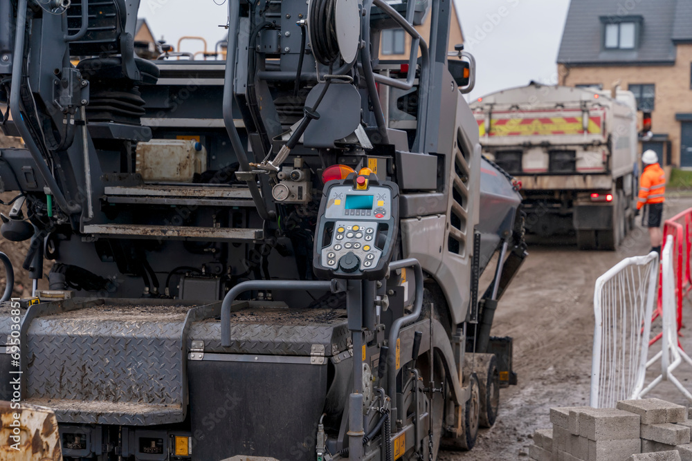 Asphalt paver filled with hot tarmac laying new road surface on new residential housing development site and roadworker operator in orange hi-viz next to it