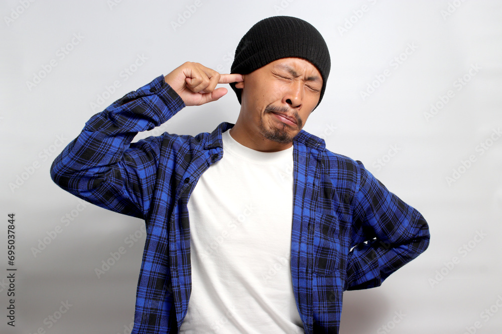 Young Asian man, wearing a beanie hat and casual shirt, is seen itching and inserting a finger into his ear, possibly due to irritation or itchiness, isolated on a white background Young Asian man, wearing a beanie hat and casual shirt, is seen itching and inserting a finger into his ear, possibly due to irritation or itchiness, isolated on a white background