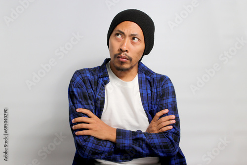 An angry, offended young Asian man stands with his arms folded, displaying an unsatisfied or discontented demeanor while looking aside at empty copy space, isolated on white background. photo