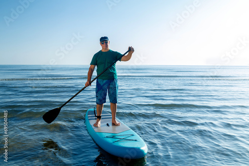 A man in shorts and a T-shirt stands on a SUP board with a paddle near the sea. photo