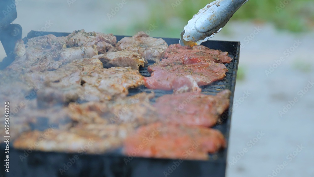 Barbecue smoke and flames, grilled meat on fire. Barbecue outdoors. Frying a raw meat steak on a burning fire, a person uses tongs. Close-up of meat steak cooked on the grill. Summer picnic concept Barbecue smoke and flames, grilled meat on fire. Barbecue outdoors. Frying a raw meat steak on a burning fire, a person uses tongs. Close-up of meat steak cooked on the grill. Summer picnic concept
