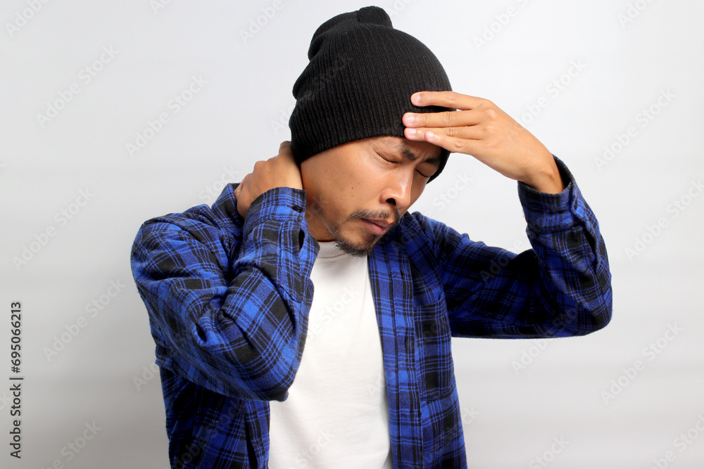 Young Asian man, dressed in a beanie hat and casual shirt, is holding his head, suggesting he's experiencing a headache, isolated on a white background. Healthcare and medical concepts Young Asian man, dressed in a beanie hat and casual shirt, is holding his head, suggesting he's experiencing a headache, isolated on a white background. Healthcare and medical concepts