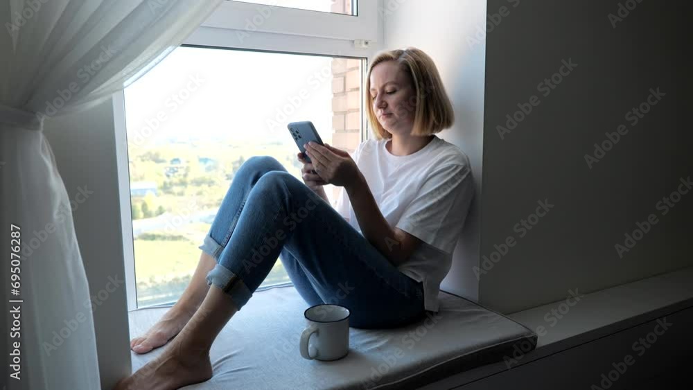 A woman sits by the window with a phone in her hands. View of a young blonde sitting by the window with a cup of hot tea. Correspondence on social networks using a smartphone A woman sits by the window with a phone in her hands. View of a young blonde sitting by the window with a cup of hot tea. Correspondence on social networks using a smartphone