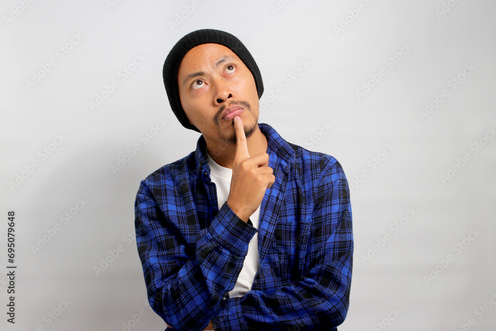 Thoughtful young Asian man, wearing a beanie hat and casual shirt, rests his hand on his chin, looking aside at empty copy space, deeply engrossed in thought and planning for the future Thoughtful young Asian man, wearing a beanie hat and casual shirt, rests his hand on his chin, looking aside at empty copy space, deeply engrossed in thought and planning for the future