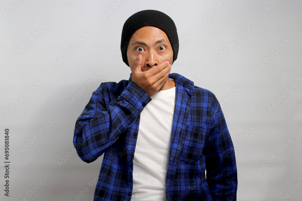 A surprised young Asian man, dressed in a casual shirt and sporting a beanie hat, is signaling for quiet by covering his mouth with his hands and shushing, while standing against white background.