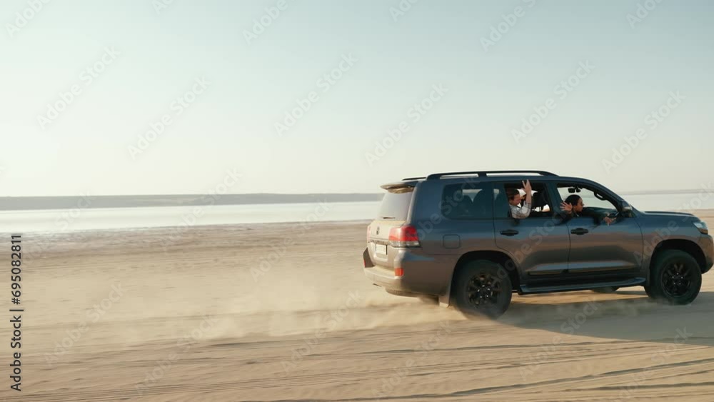 Happy multinational friends waving at camera and driving in car near estuary on sand in summertime Happy multinational friends waving at camera and driving in car near estuary on sand in summertime