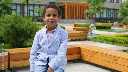 Happy boy in jacket sit on bench in park near business center photo
