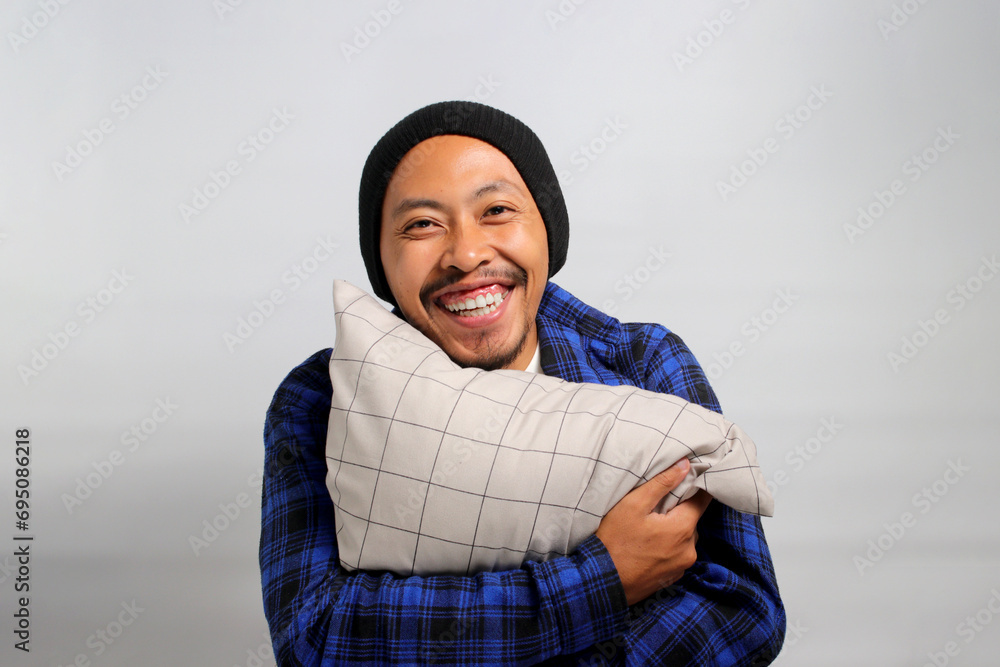 Cheerful young Asian man, dressed in a beanie hat and casual shirt, hugs a pillow, looking at the camera with a wide, toothy smile while standing against a white background Cheerful young Asian man, dressed in a beanie hat and casual shirt, hugs a pillow, looking at the camera with a wide, toothy smile while standing against a white background