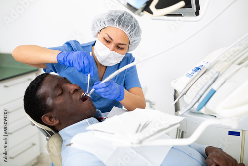 Asian woman dentist using tools for tooth repairing on african-american man patient in clinic.