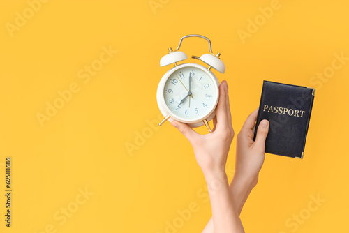 Female hands holding alarm clock and passport on yellow background photo
