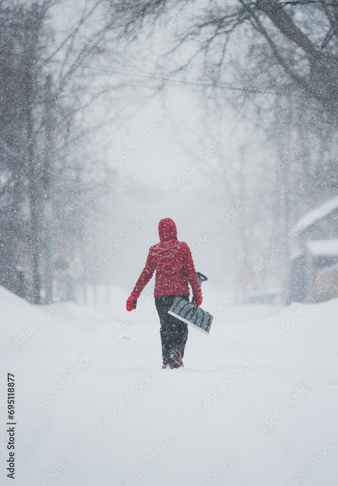 A person in a red jacket carries a shovel down a street during a snowstorm