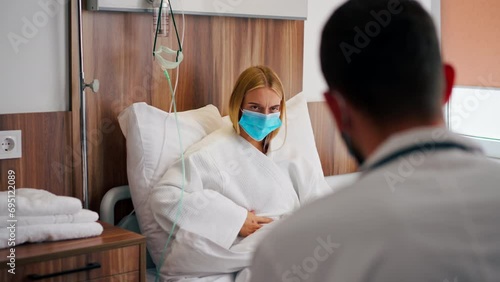 A girl in a hospital room in a medical mask holds her stomach with her hand in pain and listens to doctor with his recommendations photo