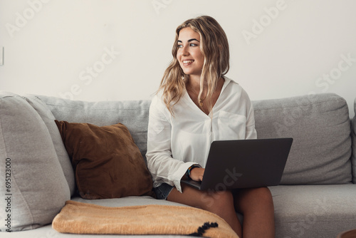 Portrait of one young attractive blonde woman using laptop pc computer on couch relaxing surfing the net at home looking at the window waiting package photo