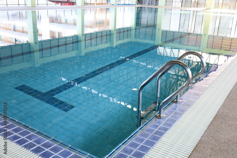 Metal stairs in the pool. Shiny stainless steel railing next to a swimming pool in a building with a roof and reflection in the water with copy space. Select focus
