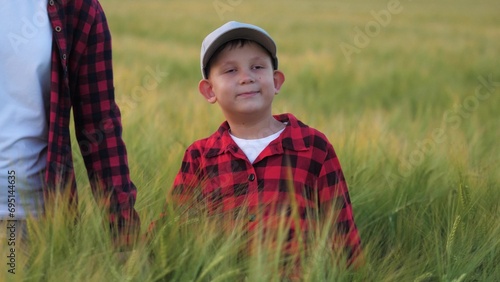 Son walks in middle of tall ears of wheat with mother on field. Kid of farmers strolls through wheat field tightly holding mother hand. Content child hand in hand with mother explores wheat field