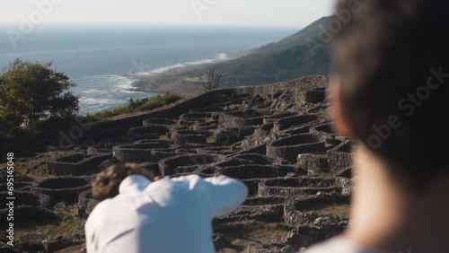 Sunlit summer eve on Santa Tecla, A Guarda. Two young men, one absorbed in the view, the other setting up a camera on a tripod. Ancient Castro, lush hills and Atlantic ocean provide a serene backdrop. photo