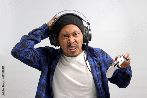 An angry and displeased young Asian man, dressed in a beanie hat and casual shirt, appears stressed while playing a video game using a gamepad on a game console, isolated on white background