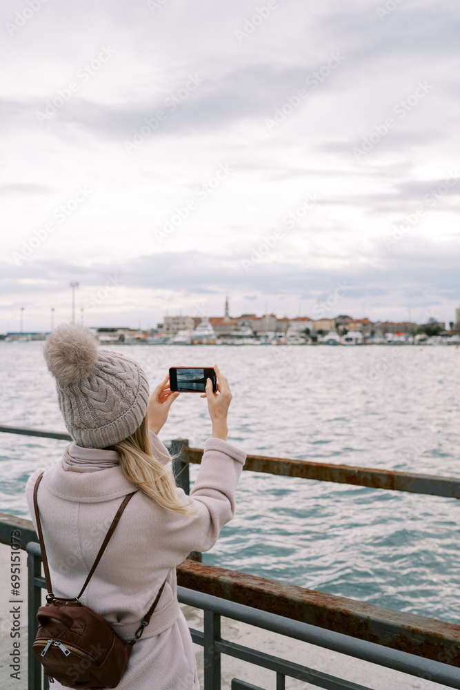 Girl with a backpack stands on the pier at the railing and takes pictures of houses on the seashore with a smartphone. Back view