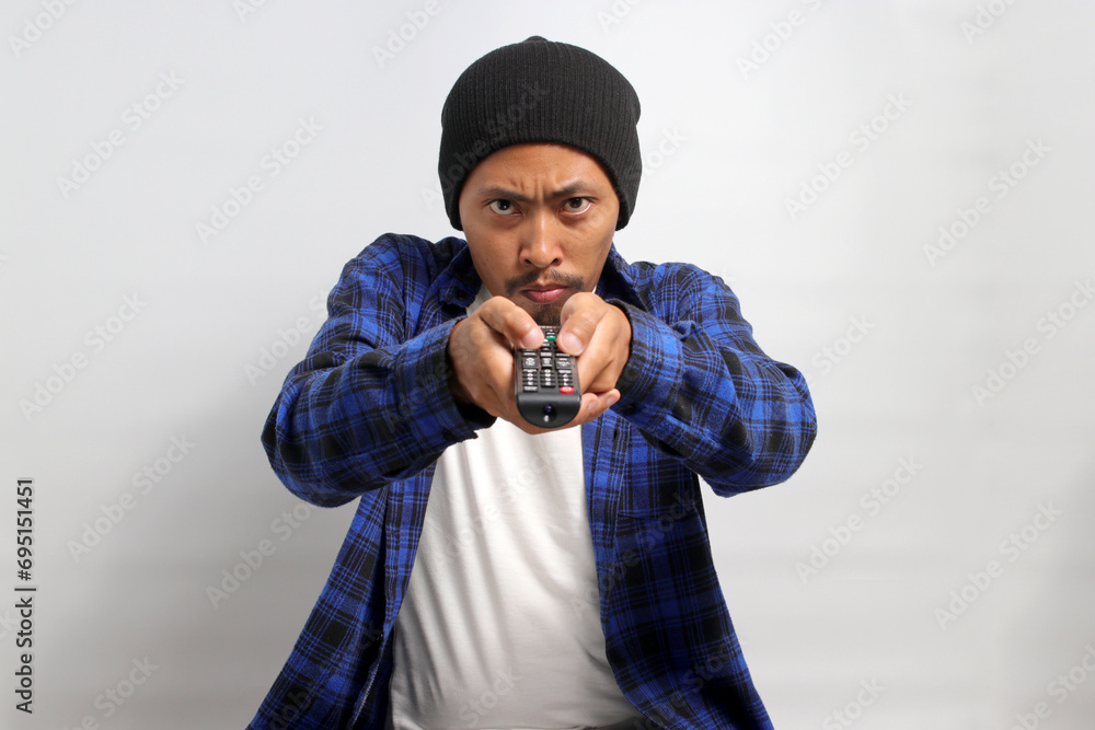 An irritated young Asian man, dressed in a beanie hat and casual shirt, switches TV channels, expressing feelings of upset and boredom, depicted against a white background. An irritated young Asian man, dressed in a beanie hat and casual shirt, switches TV channels, expressing feelings of upset and boredom, depicted against a white background.