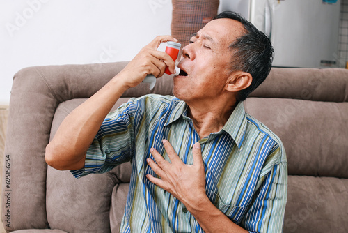 Old man sitting on couch at home holding chest having difficulties breathing and using asthma inhaler to prevent shortness of breath photo