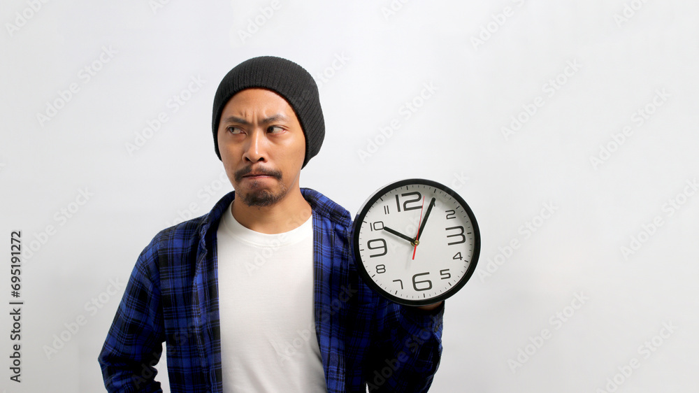 An uncertain Asian man, dressed in a beanie hat and casual shirt, holds a clock, appearing confused or doubtful as he looks aside at empty copy space while standing against a white background An uncertain Asian man, dressed in a beanie hat and casual shirt, holds a clock, appearing confused or doubtful as he looks aside at empty copy space while standing against a white background
