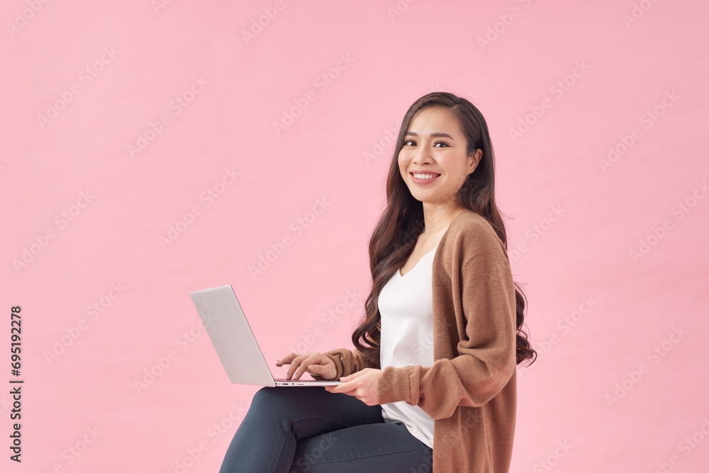 Happy young woman working online, sitting on chair and using laptop against pink wall Happy young woman working online, sitting on chair and using laptop against pink wall