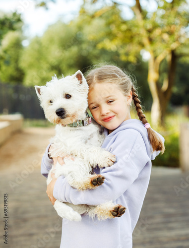 A girl with pigtails holds a small white dog in her arms and smiles happily. © Raisa Kanareva