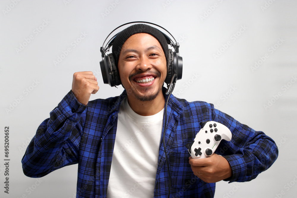 Fototapeta premium An enthusiastic Asian gamer, adorned in a beanie hat, casual shirt, and headphones, celebrates victory while playing a video game on a console with a gamepad or joystick, isolated on white background.