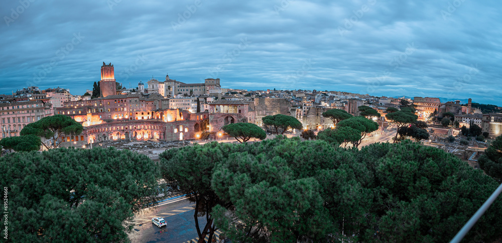 Evening view of famous rome colosseum, viewed from far. Visible road and trees leading to the famous amphitheatre, other houses and ruins visible around it.