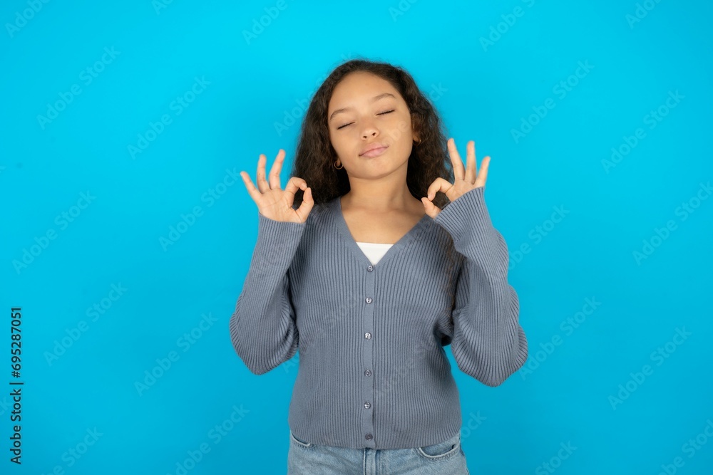 Beautiful teen girl wearing blue jacket over blue background relax and smiling with eyes closed doing meditation gesture with fingers. Yoga concept. Beautiful teen girl wearing blue jacket over blue background relax and smiling with eyes closed doing meditation gesture with fingers. Yoga concept.