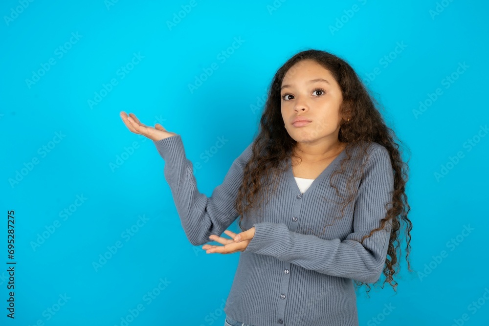 Beautiful teen girl wearing blue jacket over blue background pointing aside with both hands showing something strange and saying: I don't know what is this. Advertisement concept.