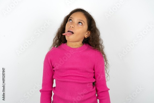 Funny Beautiful teen girl wearing pink sweater over white background makes grimace and crosses eyes plays fool has fun alone sticks out tongue.