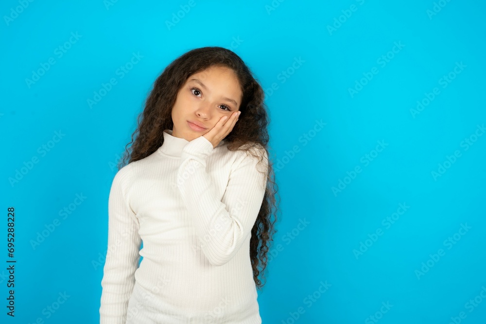 Sad lonely Young beautiful teen girl wearing white turtleneck over blue background touches cheek with hand bites lower lip and gazes with displeasure. Bad emotions Sad lonely Young beautiful teen girl wearing white turtleneck over blue background touches cheek with hand bites lower lip and gazes with displeasure. Bad emotions