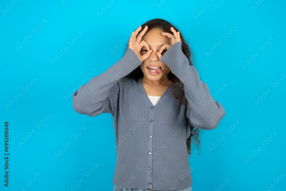 Beautiful teen girl wearing blue jacket over blue background doing ok gesture like binoculars sticking tongue out, eyes looking through fingers. Crazy expression. Beautiful teen girl wearing blue jacket over blue background doing ok gesture like binoculars sticking tongue out, eyes looking through fingers. Crazy expression.