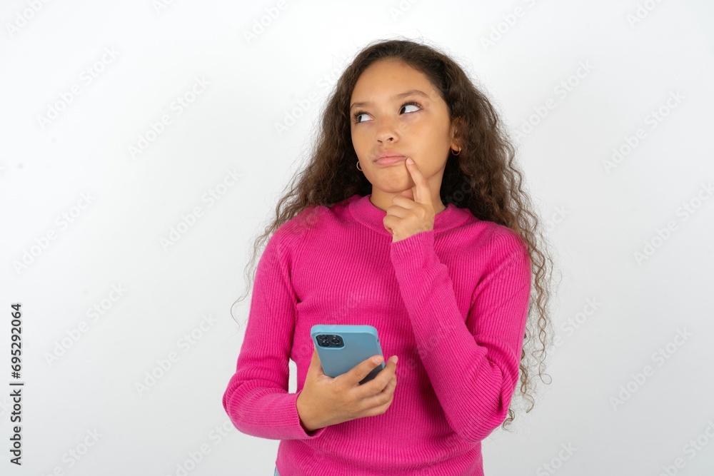 Beautiful teen girl wearing pink sweater over white background thinks deeply about something, uses modern mobile phone, tries to made up good message, keeps index finger near lips. Beautiful teen girl wearing pink sweater over white background thinks deeply about something, uses modern mobile phone, tries to made up good message, keeps index finger near lips.