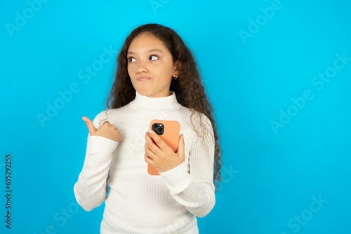 Young beautiful teen girl wearing white turtleneck over blue background points thumb away and shows blank space aside, holds mobile phone for sending text messages.